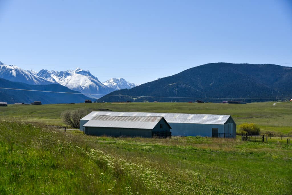 A metal barn sits in a green field on a cattle ranch, with mountains—some topped with snow—in the background under a clear blue sky.