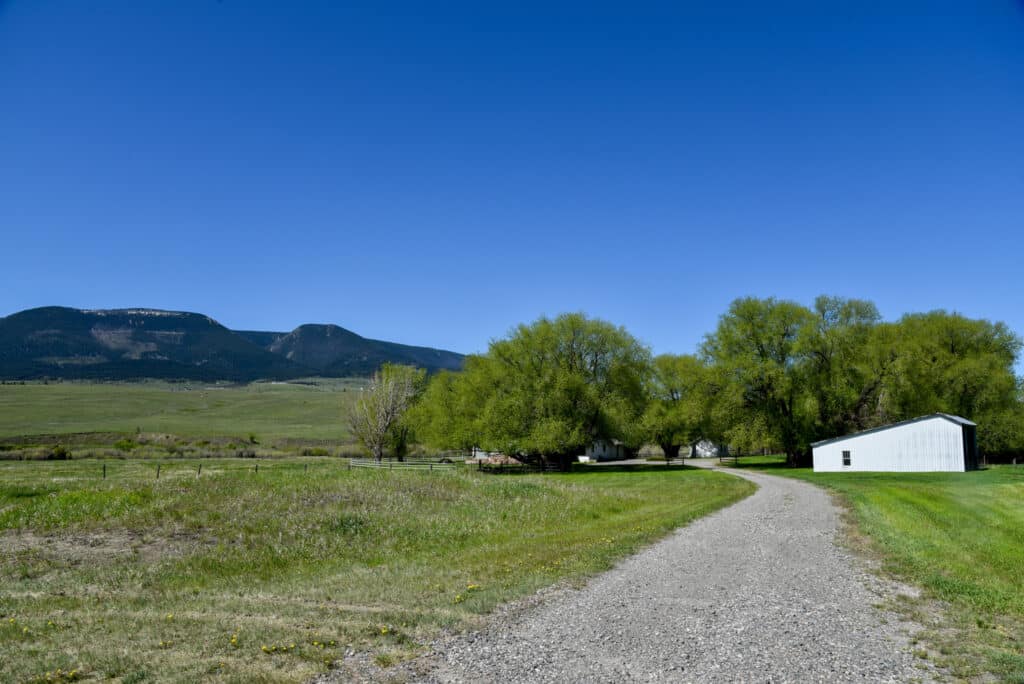 A gravel path curves through a grassy field on recreational land toward a white building, with green trees and distant mountains under a clear blue sky.