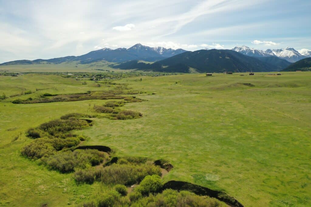 A panoramic view of a green grassy plain with a winding stream, scattered bushes, and distant snow-capped mountains under a partly cloudy blue sky—ideal land for sale perfect for a cattle ranch or hunting property.