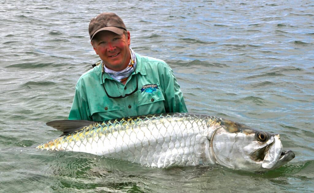 A person in a green shirt and cap stands in shallow water, smiling while holding a large silver fish with shiny scales, possibly a tarpon. The background shows rippling water—an ideal moment for anyone seeking the perfect land for sale nearby.