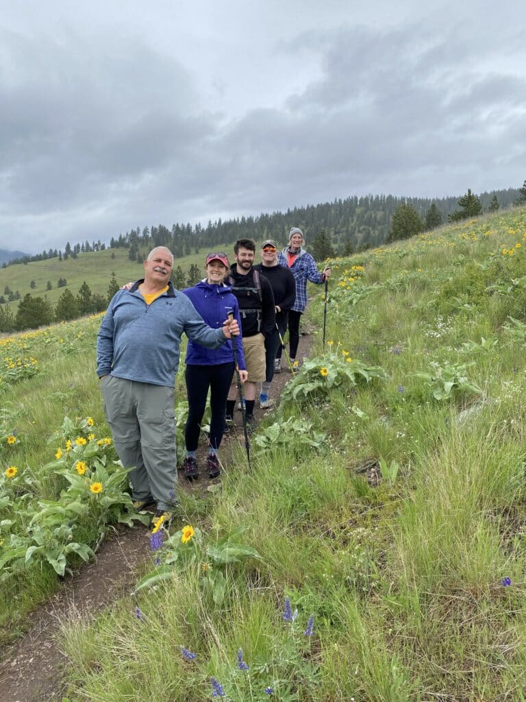 A group of six people, dressed in hiking gear and holding trekking poles, stand on a narrow trail through a green, flower-filled hillside beneath cloudy skies—perfect scenery for exploring land for sale or a scenic cattle ranch.