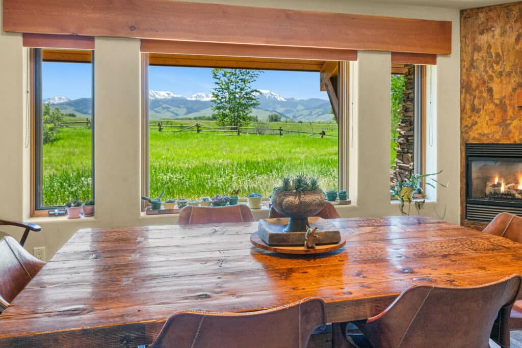 A rustic dining table and chairs sit by large windows overlooking a green field, wooden fence, and distant snow-capped mountains—perfect for enjoying a ranch for sale—with a lit fireplace nearby and small potted plants on the windowsill.