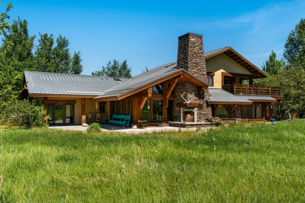 A modern rustic ranch for sale features a stone chimney, wooden beams, and a metal roof, nestled in a grassy yard surrounded by trees under a clear blue sky.
