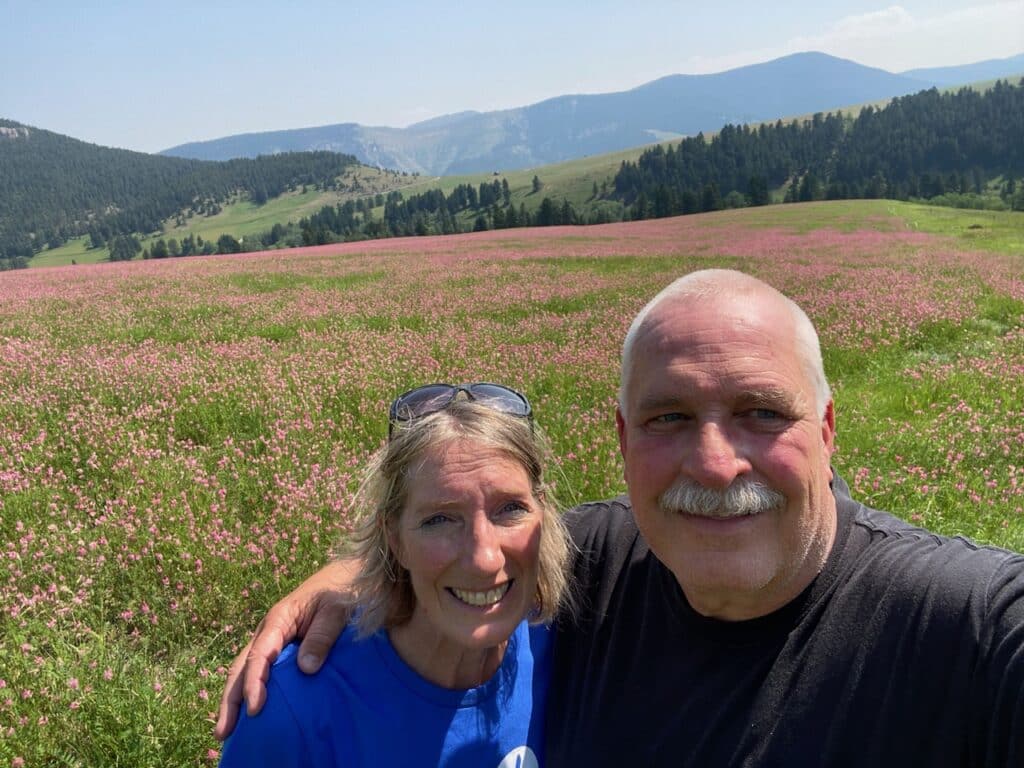 Smiling older couple takes a selfie together in front of a vast field of pink wildflowers, with rolling hills, green trees, and distant mountains under a blue sky—a perfect view for anyone seeking land for sale or dreaming of their own cattle ranch.