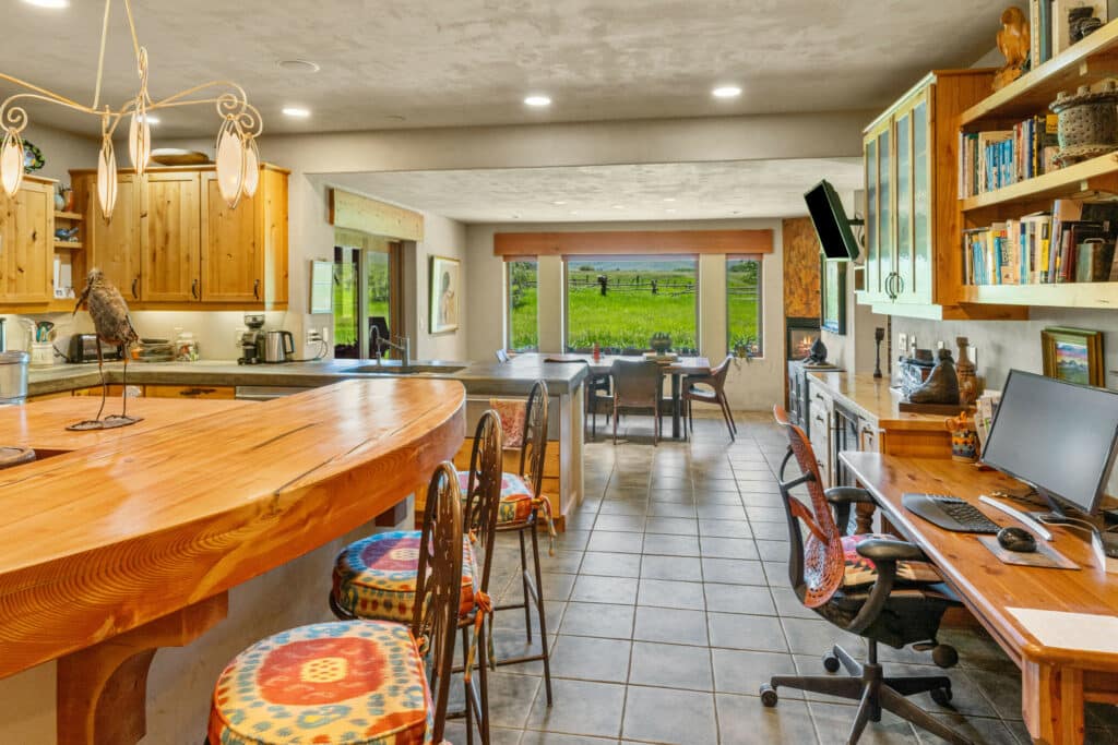 Spacious rustic kitchen and dining area with wooden cabinets, a curved wooden counter with colorful stools, tiled floor, a desk with a computer, and a large window overlooking recreational land—perfect for those seeking a unique ranch for sale.