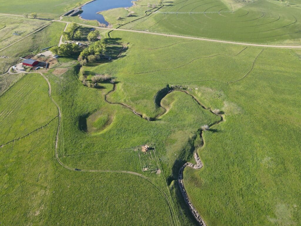 Aerial view of recreational land featuring a winding stream, grassy fields, a small pond, and a group of farm buildings surrounded by trees and fencing—ideal as a hunting property or unique land for sale opportunity.