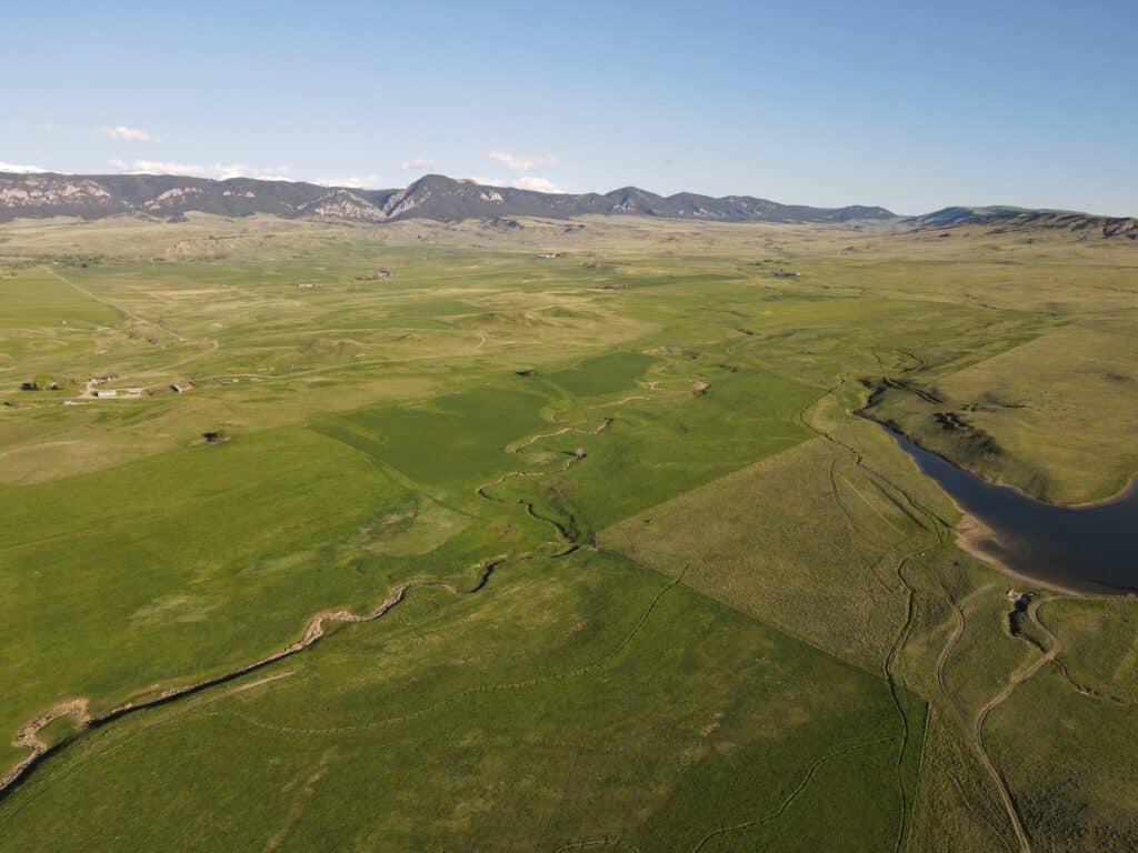 Aerial view of a vast green landscape with rolling fields, a winding stream, farmland patches, and distant mountain range under a clear blue sky—perfect setting for a ranch for sale or expansive cattle ranch.