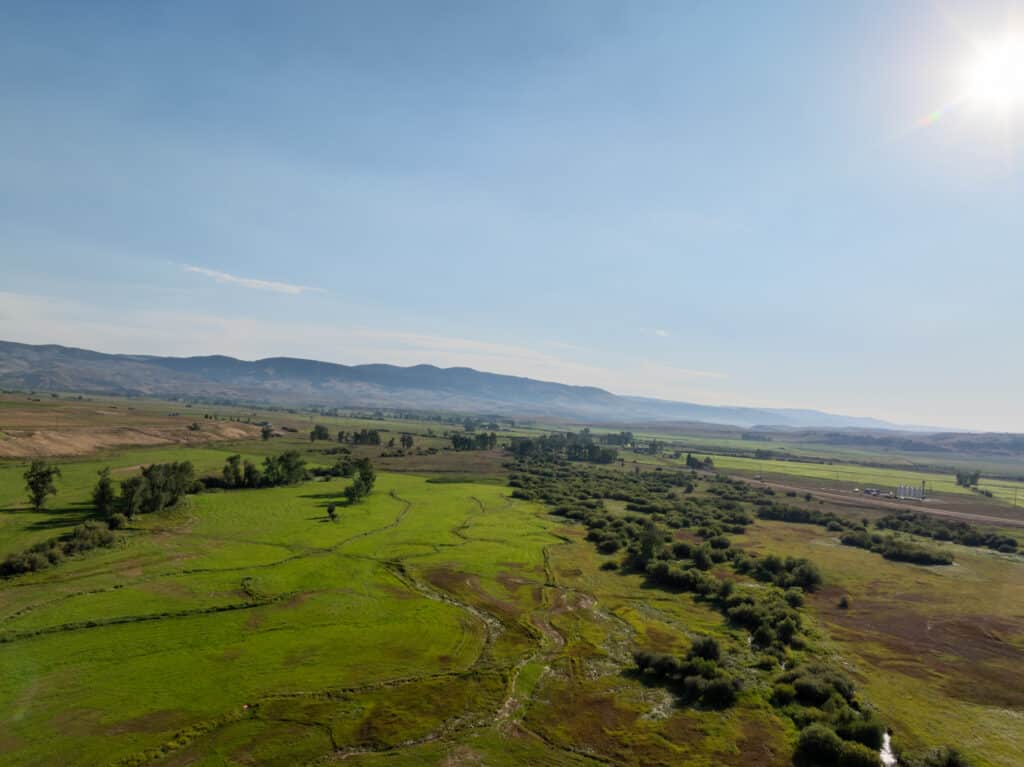 A wide view of a green valley with scattered trees and bushes, distant hills in the background, and the sun shining brightly in the clear blue sky—ideal recreational land or hunting property.