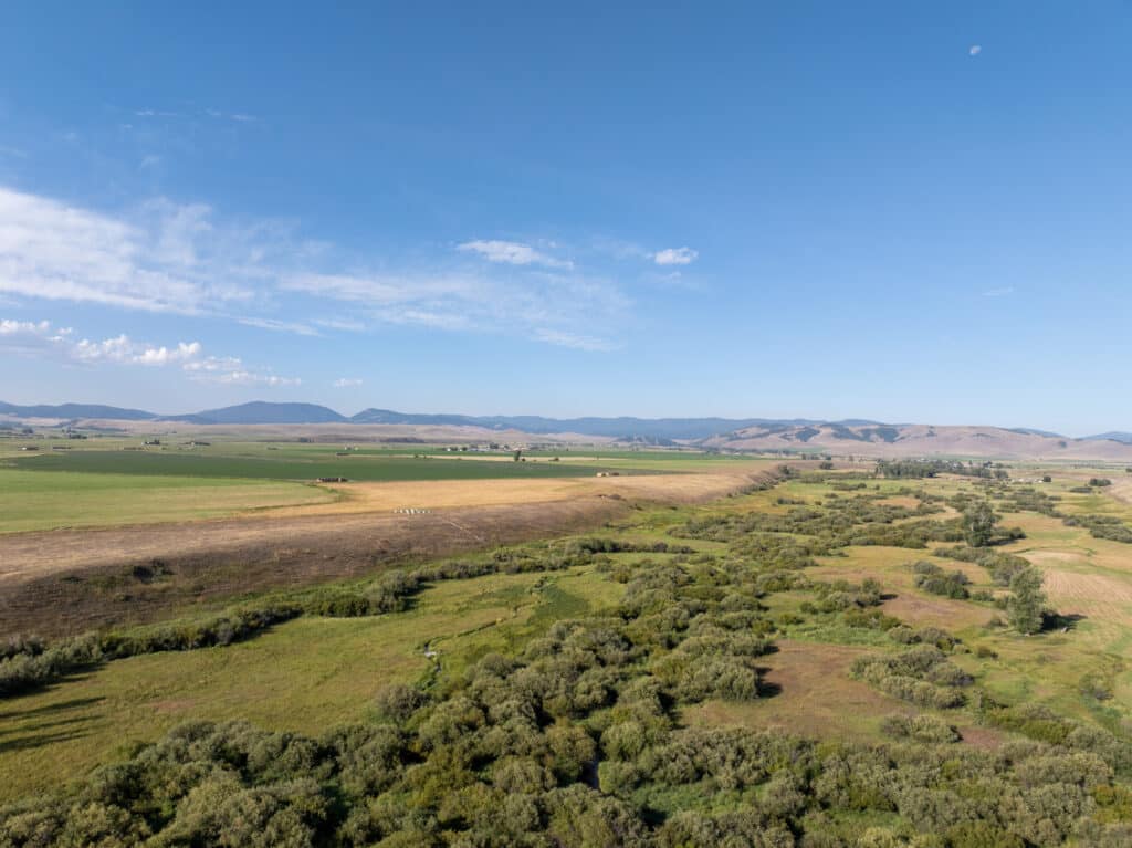 A wide view of recreational land with green fields, patches of trees, and distant hills under a clear blue sky with few clouds.