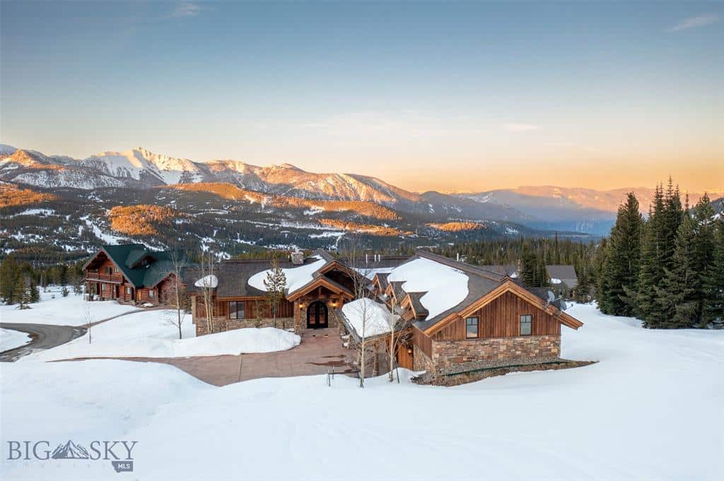 A large wooden lodge sits surrounded by snow on recreational land, with snow-covered mountains and evergreen trees in the background under a clear sky at sunset.