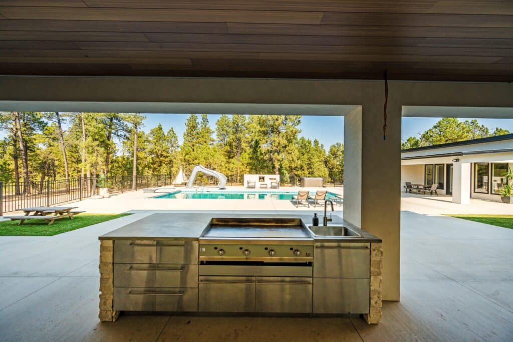 Enjoy the view from a modern outdoor kitchen with a grill and sink, overlooking recreational land with a swimming pool, poolside chairs, and lush trees under a clear sky.