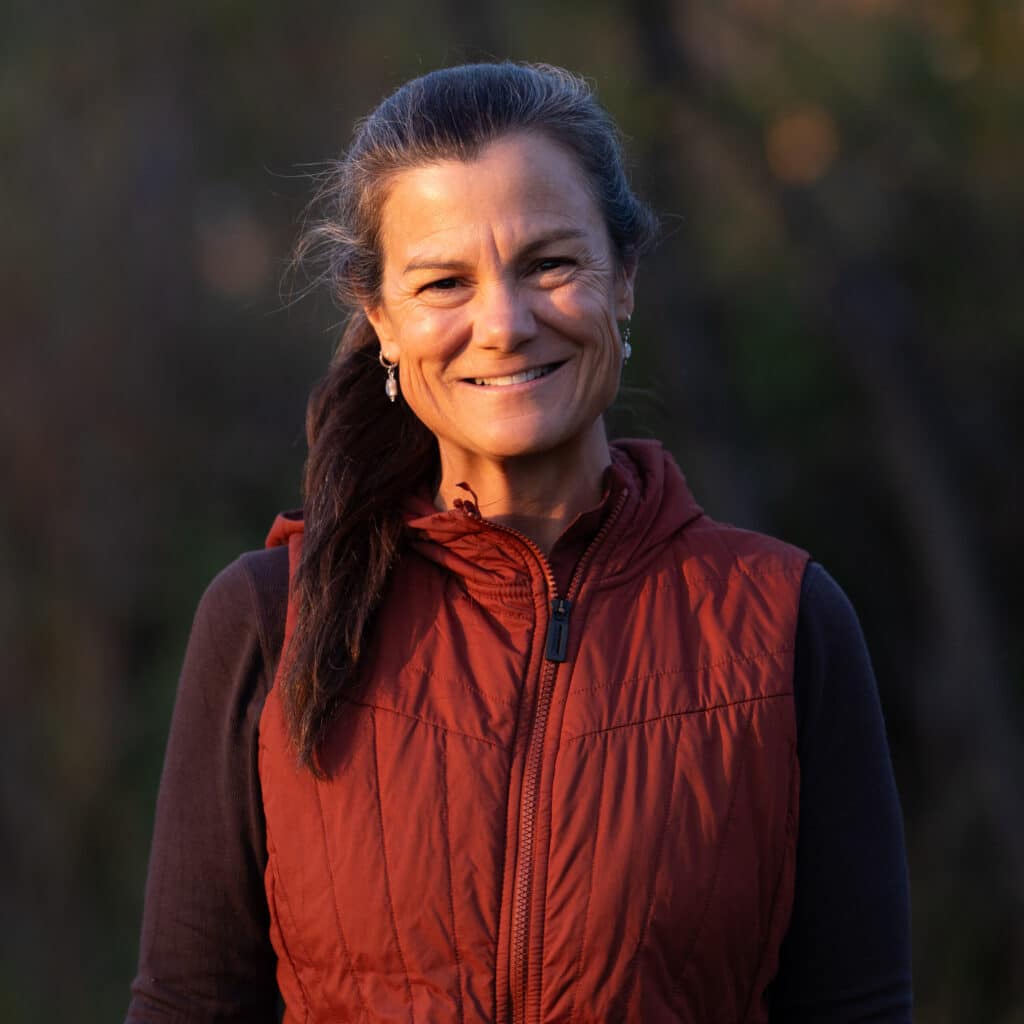 A woman with long dark hair tied back, wearing a red vest over a black top, smiles outdoors with a blurred natural background, perfect for highlighting recreational land or a ranch for sale.