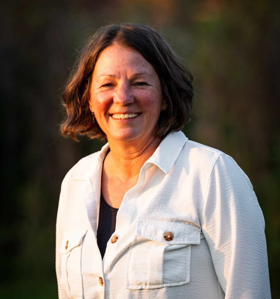 A woman with short brown hair, wearing a white button-up shirt over a dark top, smiles outdoors in warm sunlight with a blurred green background, evoking the warmth and openness of recreational land.