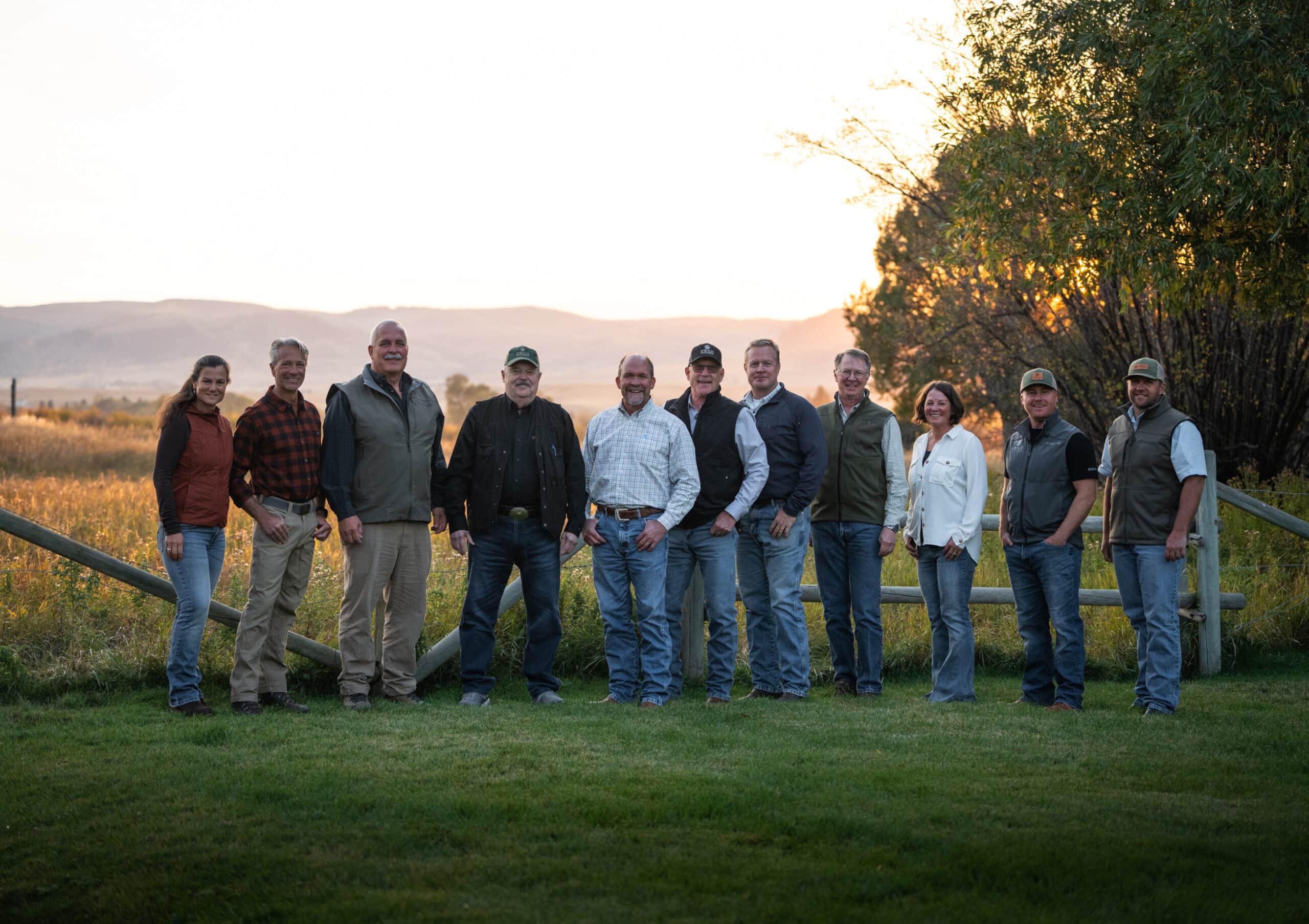A group of eleven adults stands side by side outdoors on grass, smiling at the camera, with trees and a wooden fence behind them and hills visible in the distance during sunset on a scenic recreational land.