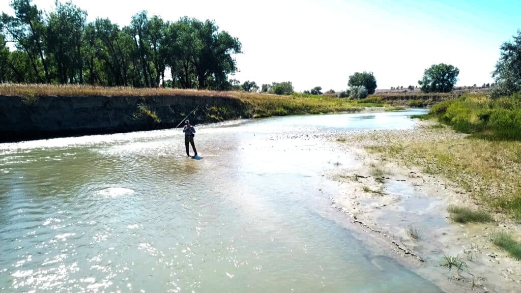 A person stands in shallow water with a fishing rod, surrounded by a riverbank with grass, mud, and trees under a bright sky—perfect scenery for exploring nearby land for sale or prime hunting property.