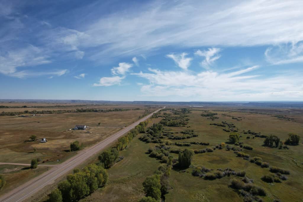 A rural landscape with a long, straight road cutting through open fields under a blue sky with wispy clouds. Scattered trees and a few small buildings hint at the potential of this recreational land or hunting property for sale.