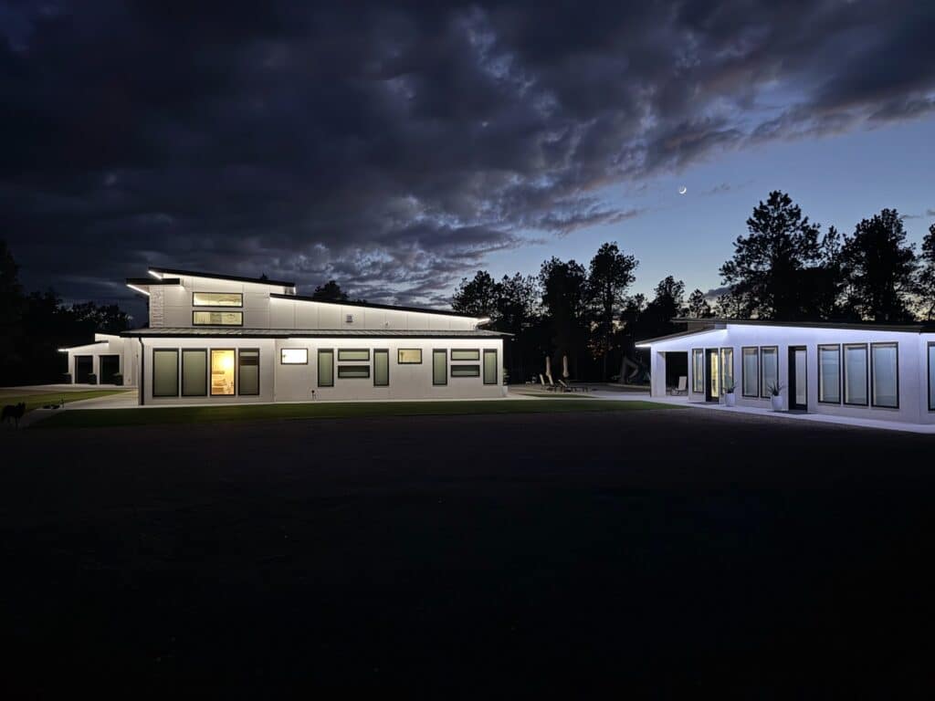 Modern buildings with large windows are illuminated at night, surrounded by trees and recreational land under a cloudy evening sky with a visible crescent moon. The area in front is dark, highlighting the buildings’ lights.