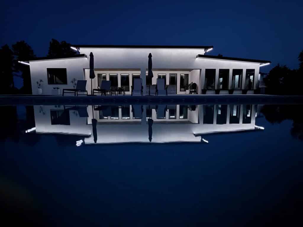 A modern white house, part of a stunning ranch for sale, is lit up at night with lights reflecting on a calm pool. The dark blue sky and outdoor furniture on the patio create an inviting, tranquil atmosphere.