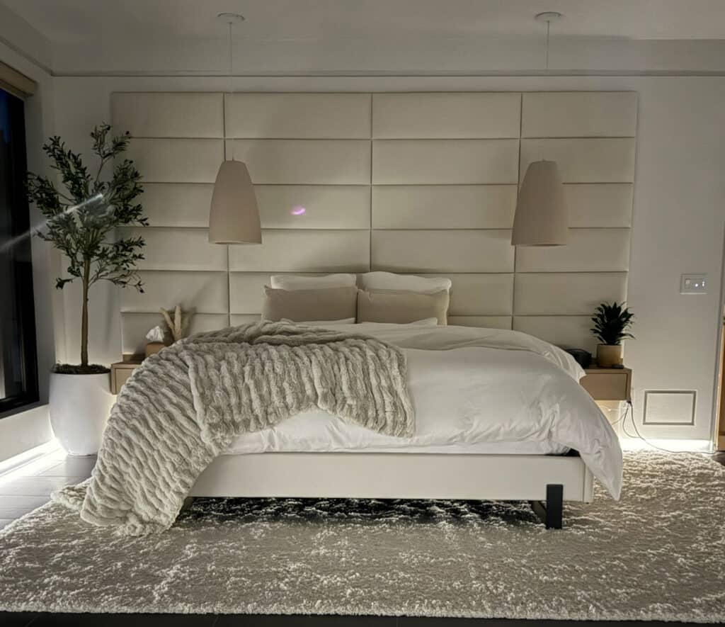 A modern bedroom with a large upholstered headboard, white bedding, a textured beige throw blanket, two pendant lights, small potted plants on nightstands, and a plush light-colored rug—perfect for unwinding after exploring ranch for sale nearby. A tall plant stands by the window.