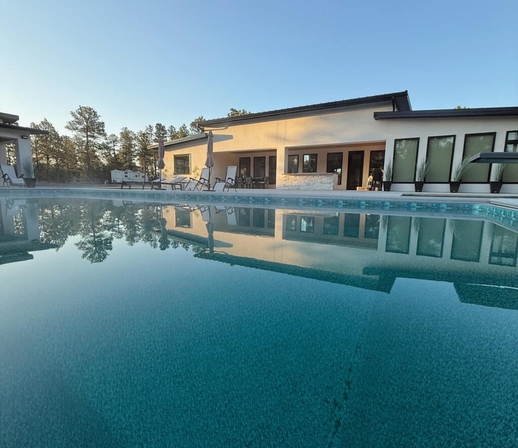 A clear outdoor swimming pool reflects a modern white building with large windows, patio umbrellas, and lounge chairs under a blue sky with surrounding trees—perfect for relaxing after a day exploring your ranch for sale.
