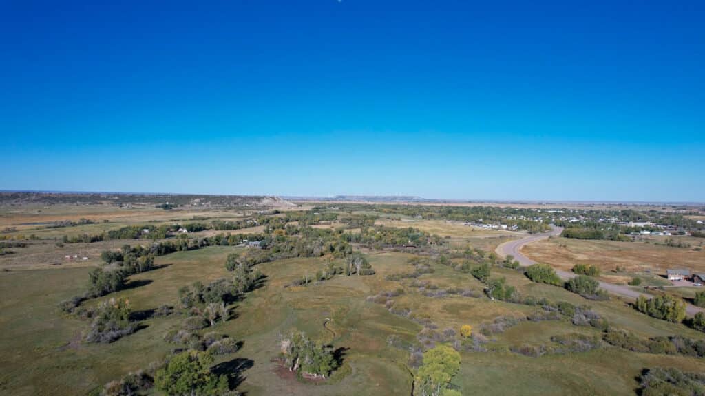 A wide aerial view of recreational land with open fields, scattered trees, a curving road, and distant houses under a clear blue sky.