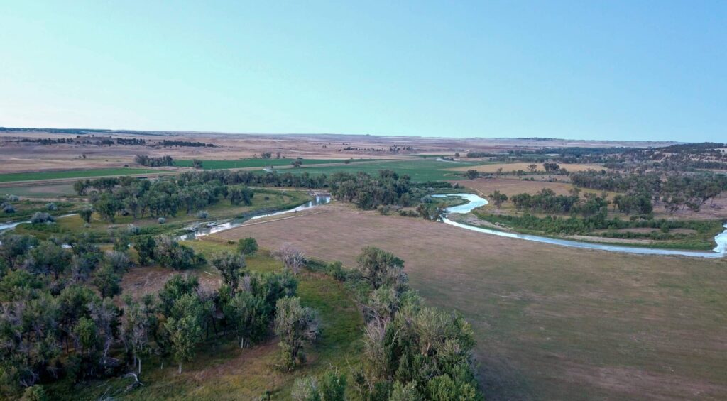A winding river flows through a flat, open landscape with patches of green trees and grass under a clear blue sky—ideal recreational land or ranch for sale amid expansive rural fields and sparse vegetation.
