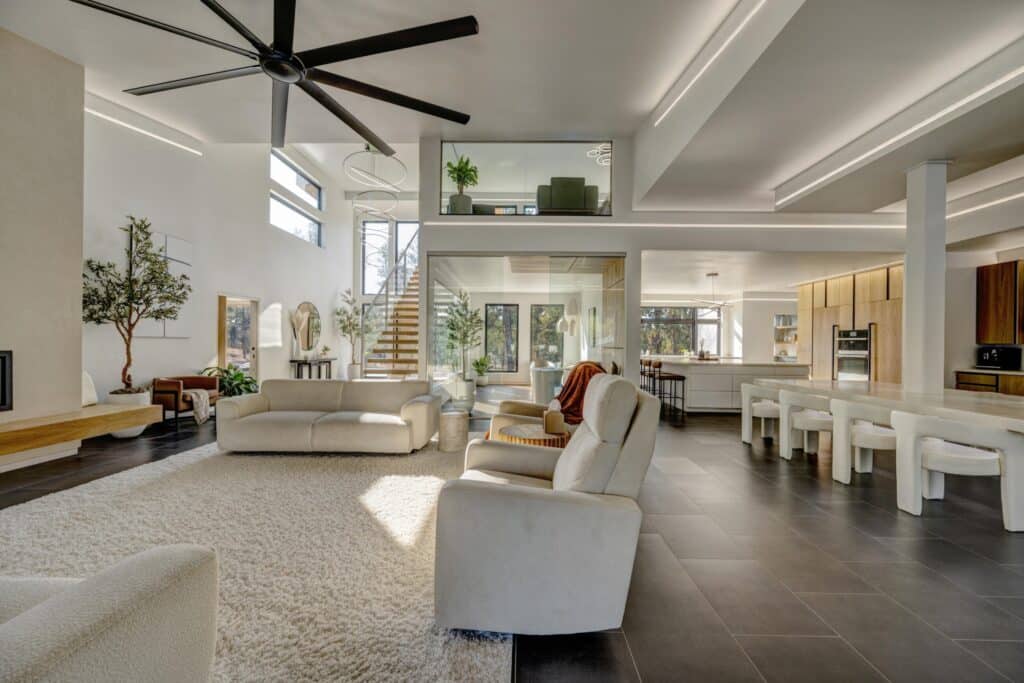 Modern open-concept living space with neutral furnishings, a large ceiling fan, kitchen with bar stools, and staircase leading to a glass-enclosed loft. Large windows let in natural light—perfect for those seeking land for sale and a peaceful retreat.