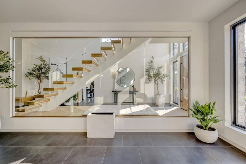 Modern interior with a large glass wall revealing a wooden floating staircase, potted plants, a round mirror, and a console table in a bright, airy space—perfect for showcasing the serene style of your hunting property or ranch for sale.