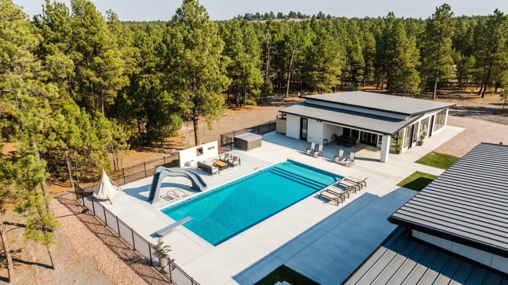 A modern house with a large backyard swimming pool, lounge chairs, and outdoor seating area sits on a picturesque cattle ranch for sale, surrounded by tall pine trees and a beautiful forested landscape.