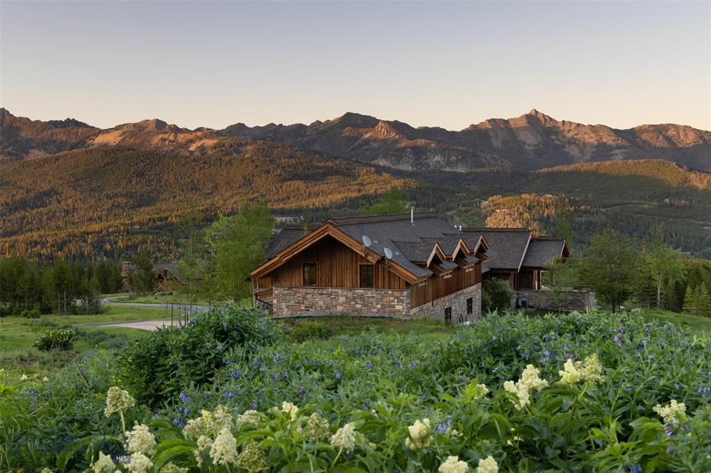 A rustic wooden house with stone accents sits among green grass and wildflowers, surrounded by trees and mountains under a clear sky at sunrise—a perfect ranch for sale amidst natural beauty.