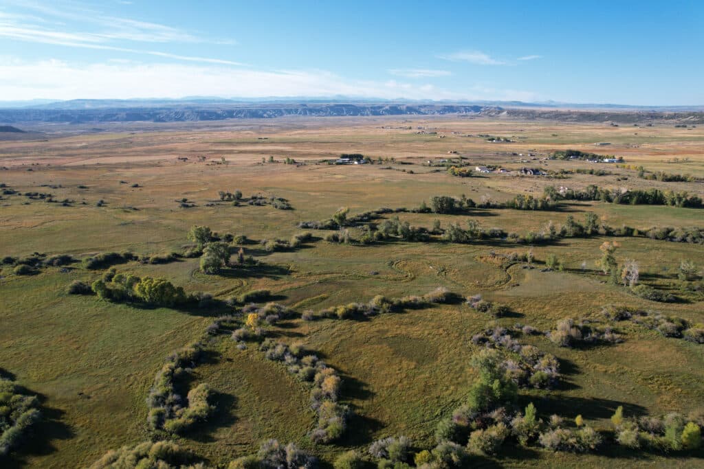 Aerial view of a vast, open grassland with patches of green trees and shrubs, distant buildings, and a backdrop of low mountains under a clear blue sky—ideal for a cattle ranch or hunting property.