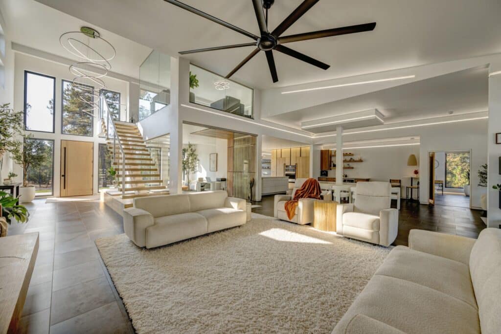 Spacious modern living room with beige sofas, a large shaggy rug, high ceilings, an oversized black ceiling fan, open staircase, and floor-to-ceiling windows. Open-concept design with kitchen and dining area ideal for a cattle ranch or recreational land setting.