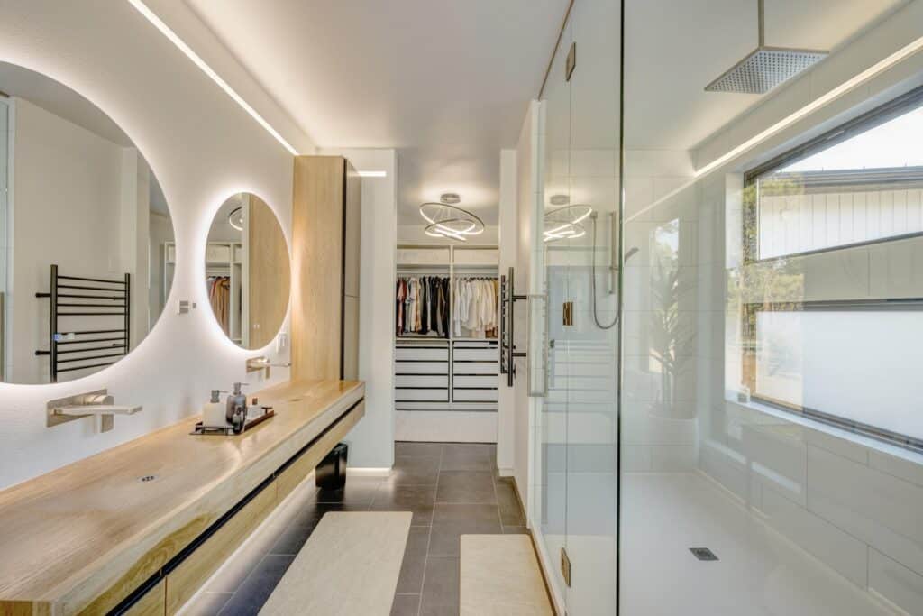 Modern bathroom with a long wooden vanity, two large round mirrors, glass shower, and a view into a walk-in closet with shelves and hanging clothes. The well-lit space offers potential inspiration for your dream home on land for sale.