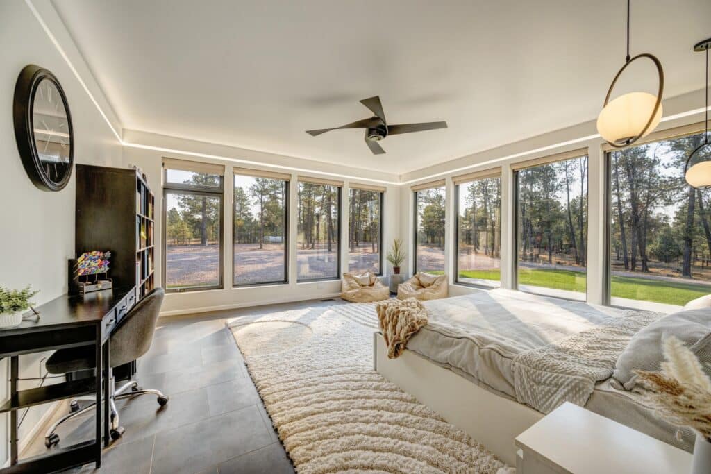Bright bedroom with large floor-to-ceiling windows overlooking trees, a cozy bed with blankets, a desk and chair, bookshelves, a ceiling fan, and bean bags on a light rug. Natural light fills this inviting space on your dream cattle ranch.