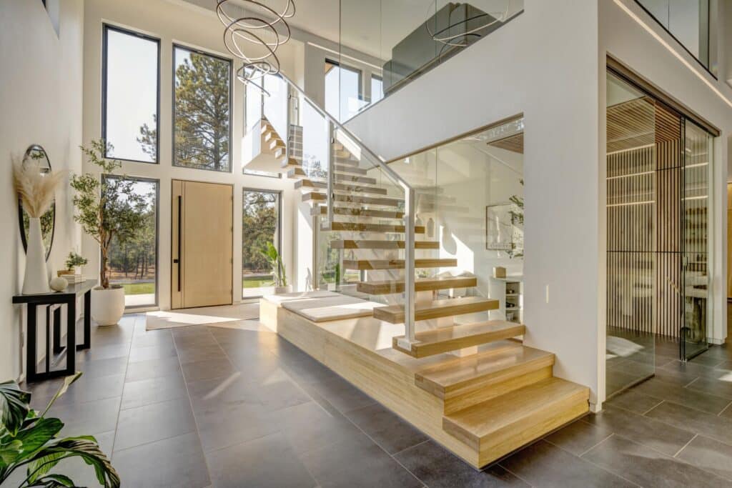 A modern home entryway with large windows and a wooden floating staircase with glass railings welcomes you, offering natural light and minimalist decor—perfect for a stylish residence on hunting property or land for sale.