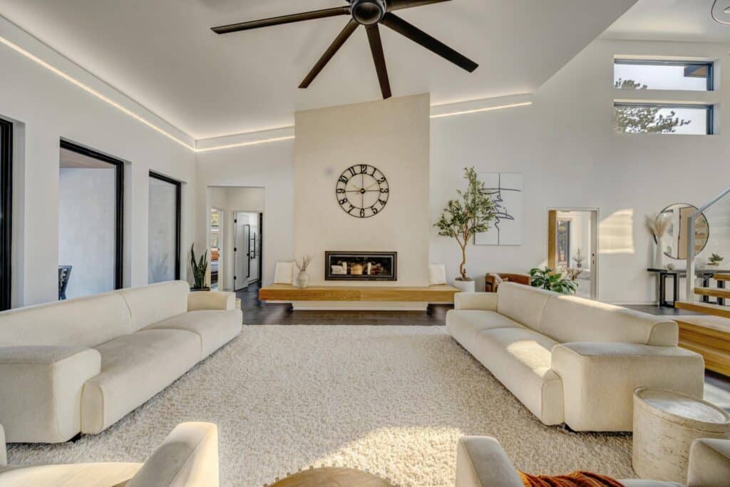 Modern living room with two cream-colored sofas, a shaggy white rug, a fireplace with a large clock above, ceiling fan, potted plants, and abundant natural light—perfect for relaxing after a day exploring your ranch for sale or recreational land.