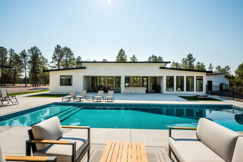 Modern white house with large windows sits behind a clear blue swimming pool. Poolside lounge chairs and cushioned patio furniture are arranged on the concrete deck. This stunning ranch for sale is framed by tall pine trees under a clear sky.