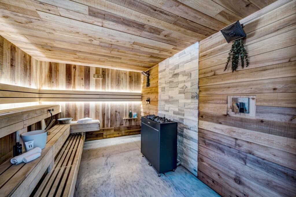 A modern wooden sauna room with benches, soft indirect lighting, a bucket and ladle, towels, and a black heater on a concrete floor beside a light stone accent wall—ideal for relaxation after enjoying recreational land.