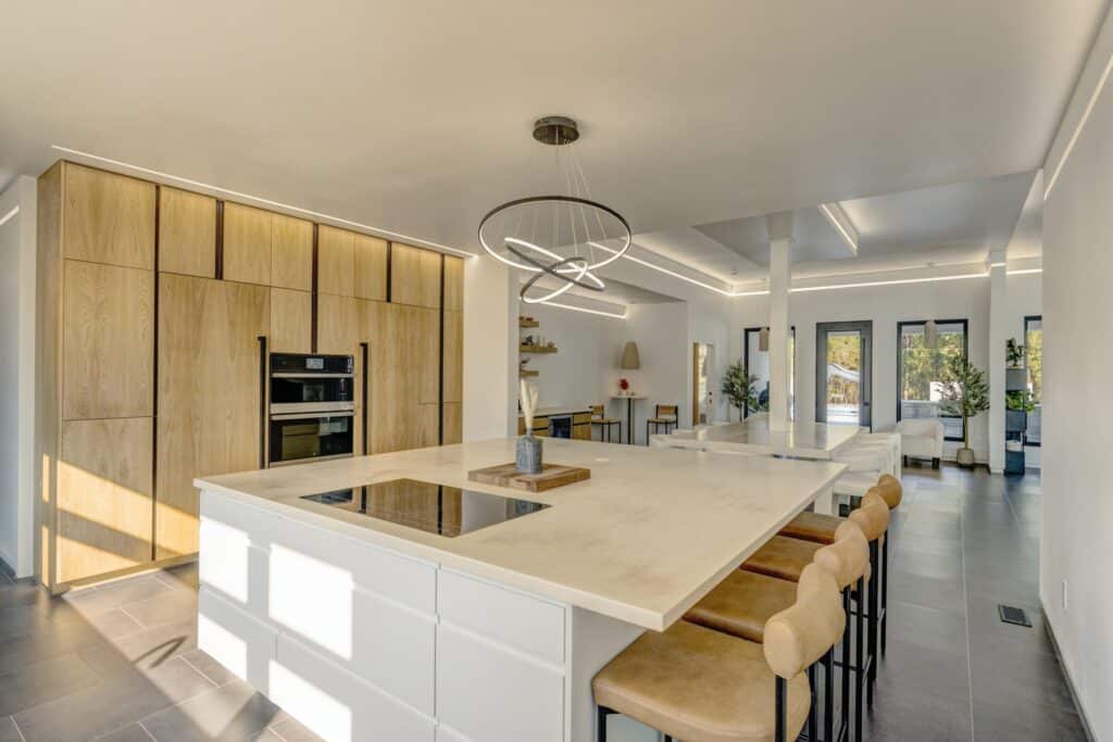 Modern kitchen with a large white island, built-in stove, beige cushioned bar stools, wood-paneled cabinets, built-in oven, minimalist decor, and ample natural light from large windows—ideal for a cattle ranch or land for sale.