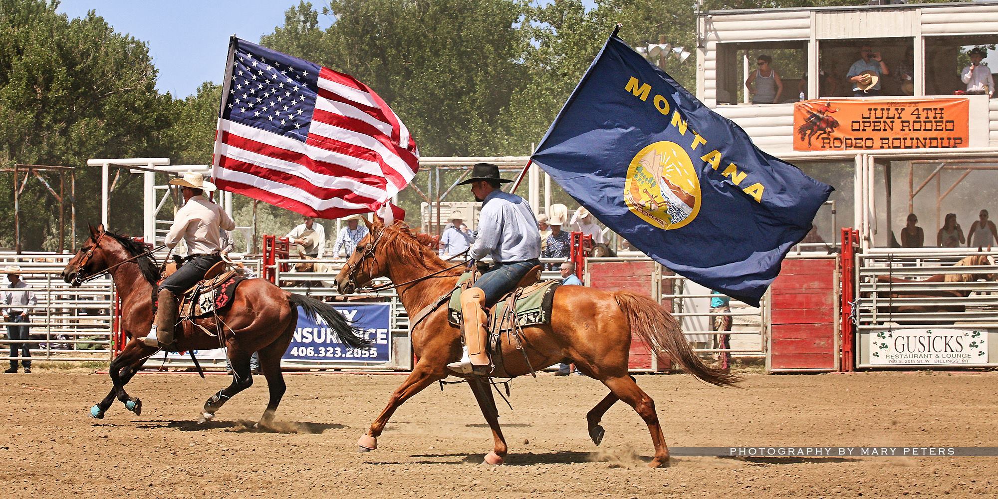 Two horseback riders in a rodeo arena carry large flags—one American and one Montana state flag. Spectators watch from bleachers, with banners in the background at this classic gathering on prized cattle ranch and hunting property land for sale.