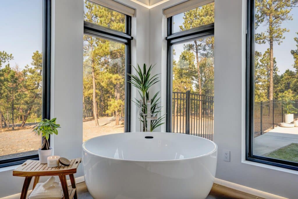 A modern bathroom with a large white freestanding bathtub by two tall corner windows showing views of trees, perfect for relaxing after a day on your cattle ranch. A wooden stool holds a candle, soap, and plant; a tall green plant stands behind the tub.