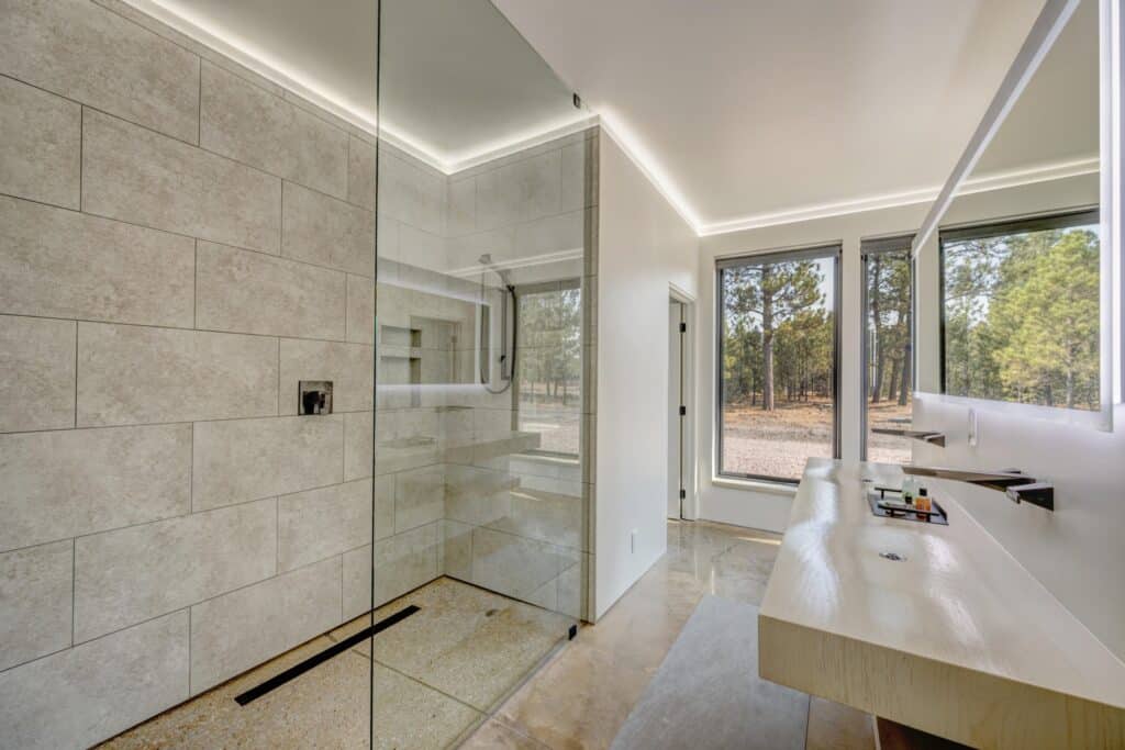 Modern bathroom with a large glass shower, beige tiles, floating vanity, wide mirror, and floor-to-ceiling windows overlooking recreational land or ranch for sale. The space is bright and minimalistic.