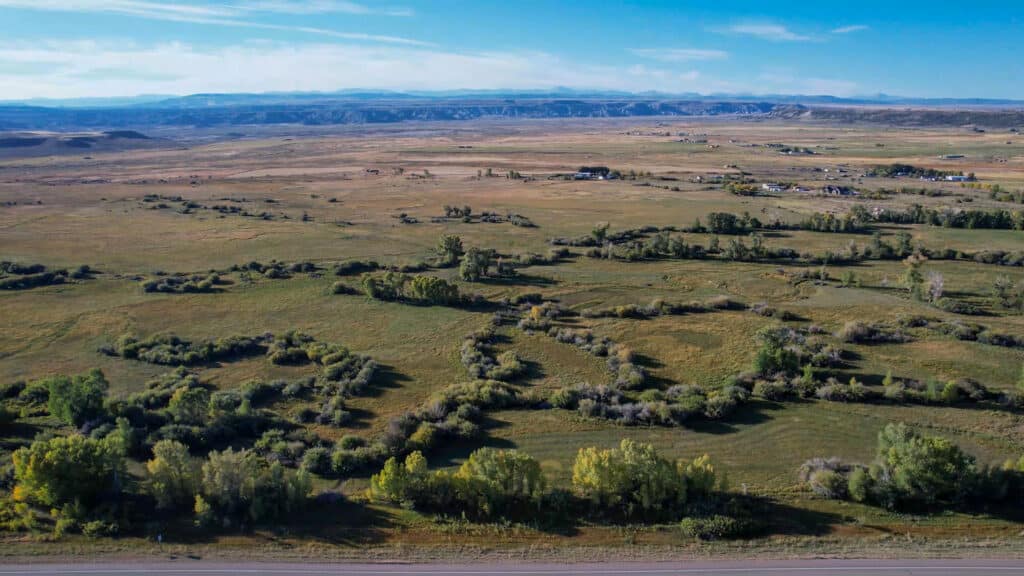 A wide aerial view of a grassy plain with a winding, tree-lined stream, scattered trees, distant houses, and mountains on the horizon under a blue sky—an ideal setting for recreational land or hunting property.