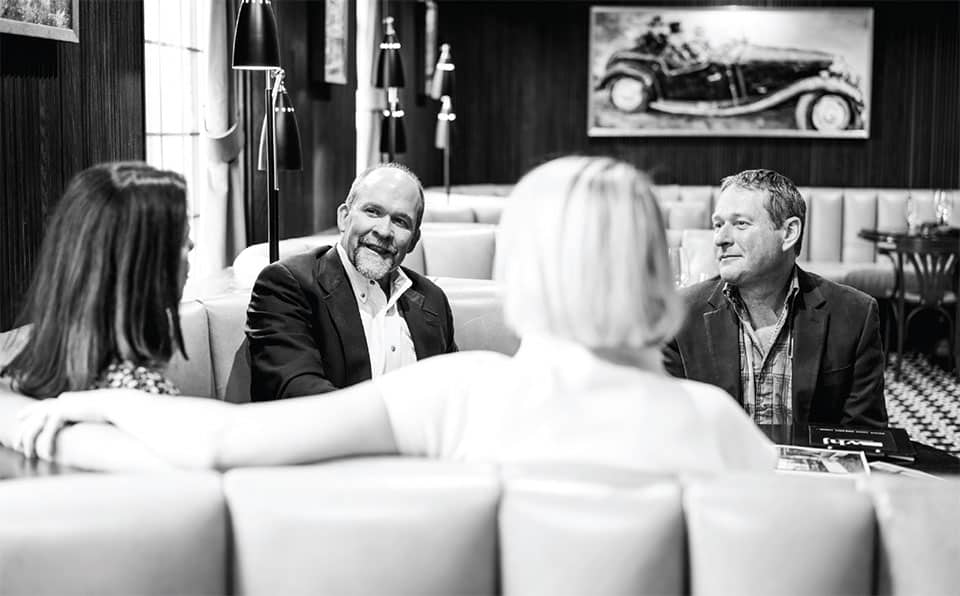 Four people sit together in a stylish restaurant booth, engaged in conversation. Two men face the camera, smiling, while two women sit with their backs to the camera. A vintage car photo hangs on the wall behind them—perfect spot to discuss hunting property or land for sale.