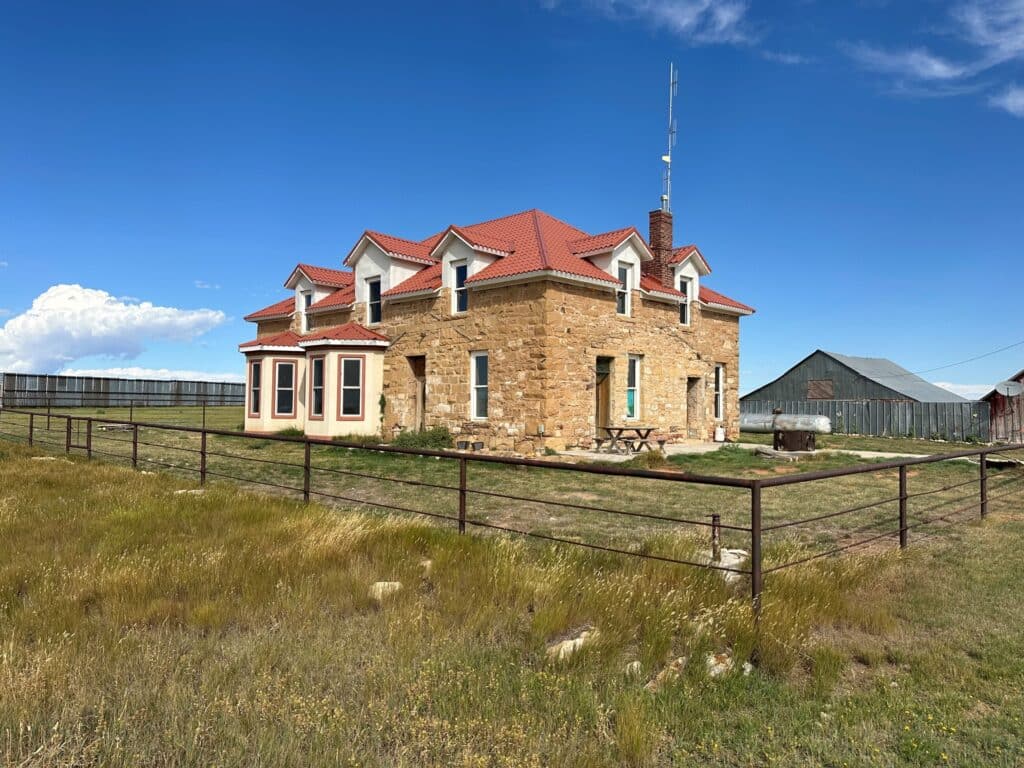 A large stone house with a red roof sits on recreational land in a grassy fenced yard under a blue sky. An old barn and wooden fence are visible in the background. The house features dormer windows and a small bay area extension.