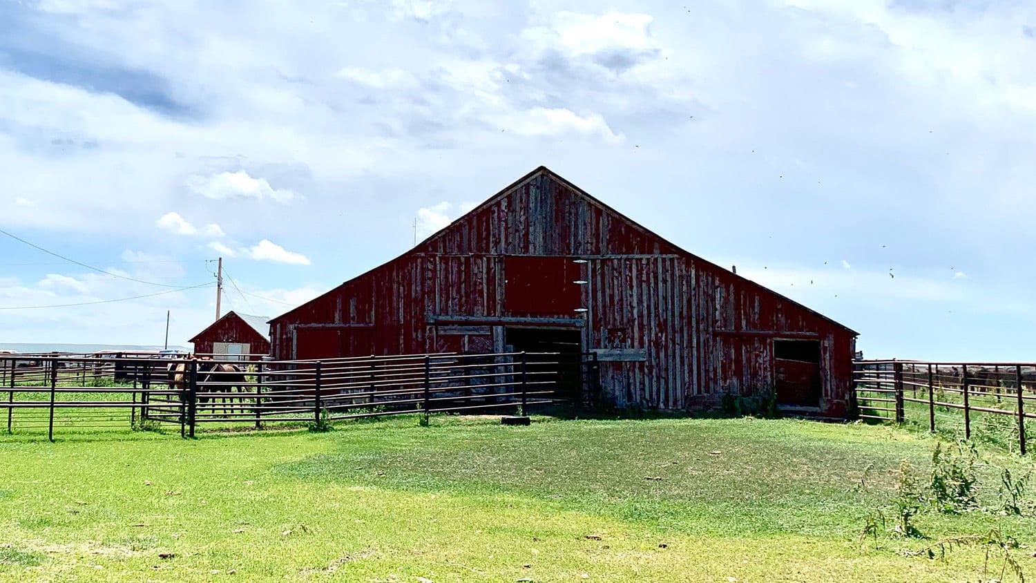 A weathered red barn stands in a grassy field under a partly cloudy sky, surrounded by metal fences, making this ranch for sale perfect for recreational land use. Another small barn is visible in the background.