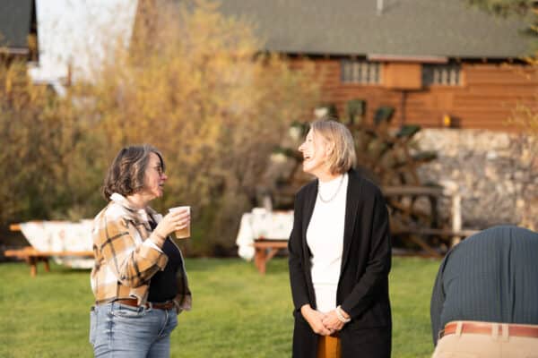 Two women stand outside on the grass of a cattle ranch, smiling and talking. One holds a drink in a plaid jacket; the other wears a black sweater and white top. A man stands nearby, bent over, with a rustic building in the background.