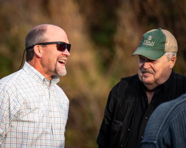 Two men stand outdoors on recreational land, one in sunglasses and a plaid shirt smiling, the other in a green cap and black jacket, both engaged in conversation with a blurred background.