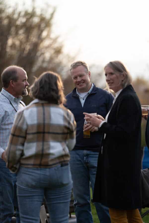 Four adults stand outdoors on recreational land, talking and smiling. One man holds a drink while another faces a woman in a plaid shirt. Another woman in a black coat stands in the foreground, with blurred trees in the background.