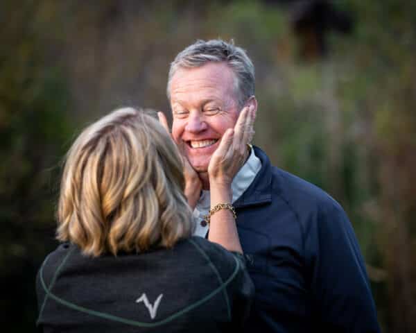 A woman gently holds a smiling man's face in her hands outdoors, both appearing happy and playful. The blurred greenery hints at open recreational land, creating a relaxed and joyful atmosphere.